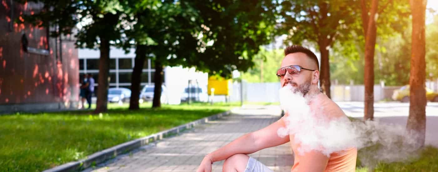 A young man with sunglasses is vaping on the sidewalk.